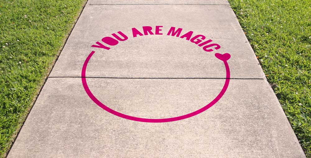 Chalk art on sidewalk surrounded by green grass reads "You Are Magic" in pink text