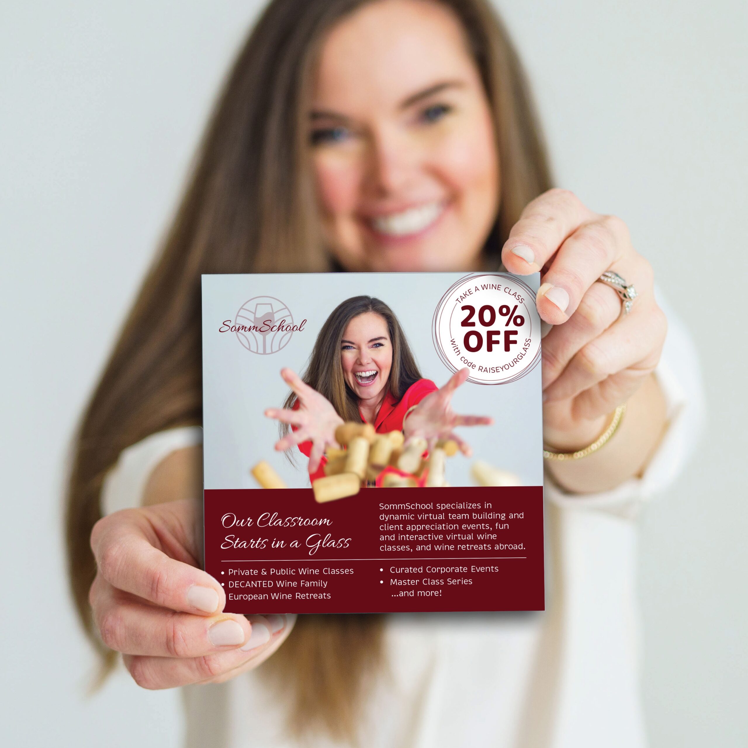 A square promotional flyer shows a smiling woman throwing corks towards the camera. A woman with long brown hair wearing white is holding up the card and standing blurred out in the background.
