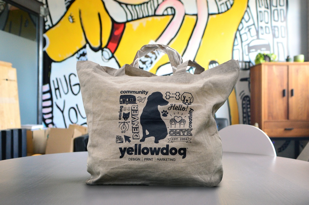 Tan tote bag sitting on a table in front of a large yellow dog mural with black YellowDog branding