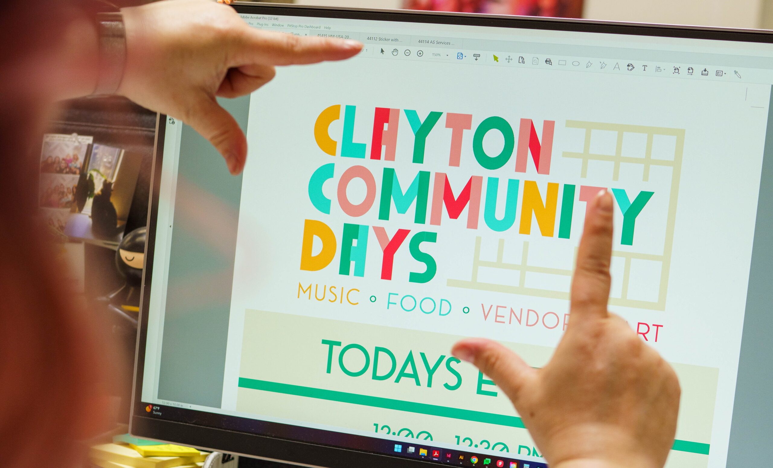A YellowDog-designed event promotion poster is shown on a computer screen framed by two hands. The colorful poster reads: "Clayton Community Days, Music, Food, Vendors, Art, Today's ...".