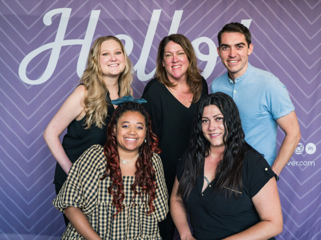 A group of five people poses in front of a purple banner that reads 'Hello!'