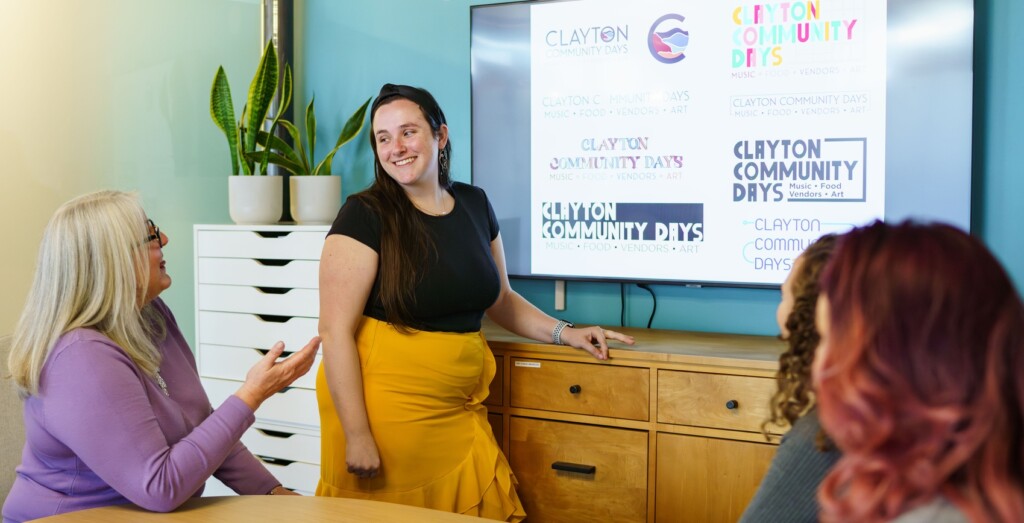 Workshopping design--A woman is pictured standing in front of a large television screen with eight different logo designs on it. She is looking at a woman sitting to her right who is talking and gesturing toward the screen.